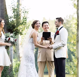 Bride and groom holding hands and smiling during outdoor wedding ceremony officiated by a man in beige suit.