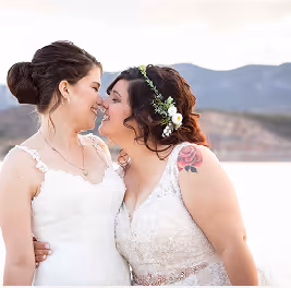 Two brides in white wedding dresses smiling and embracing outdoors with mountains in the background.