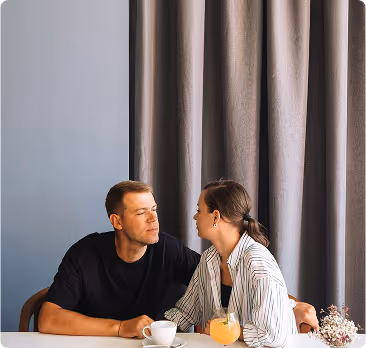 A man and woman sitting at a table holding hands, with a coffee cup and a glass of orange juice in front of them, against a backdrop of gray curtains.