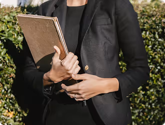 Person in a black blazer holding three notebooks outdoors with greenery in the background.