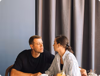 A couple sitting closely and looking at each other indoors with curtains in the background.
