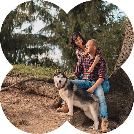 Couple sitting on a tree root outdoors with a Siberian Husky dog standing in front of them.