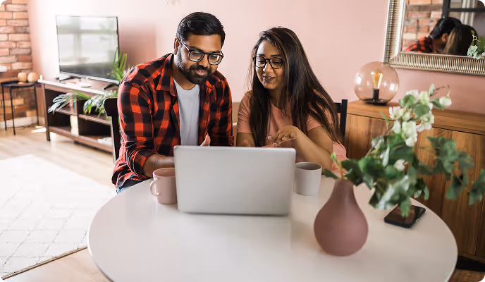 A man and woman with glasses sitting at a round table using a laptop together, with mugs and a flower vase nearby.