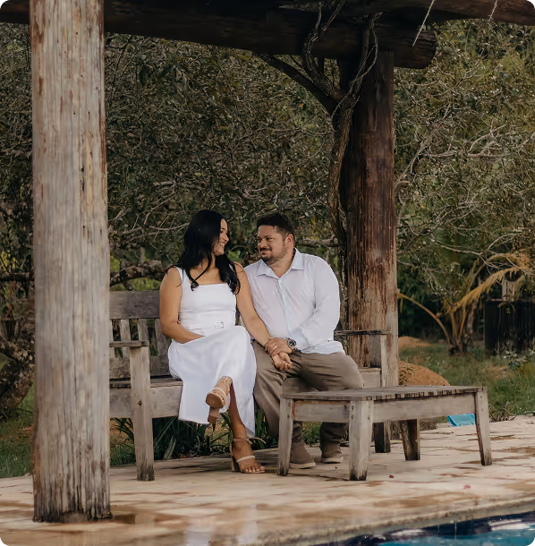 A couple sitting hand-in-hand on a wooden bench under a rustic shelter near a pool, surrounded by trees.