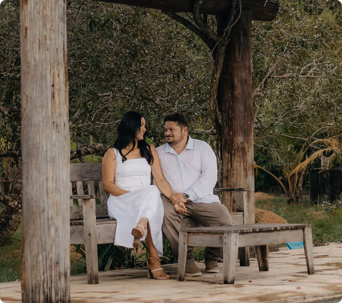 Couple sitting on a wooden bench under a pergola, holding hands and looking at each other, surrounded by greenery.