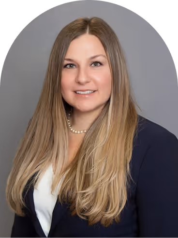 Portrait of a woman with long blonde hair wearing a navy blazer, white blouse, and pearl necklace against a gray background.