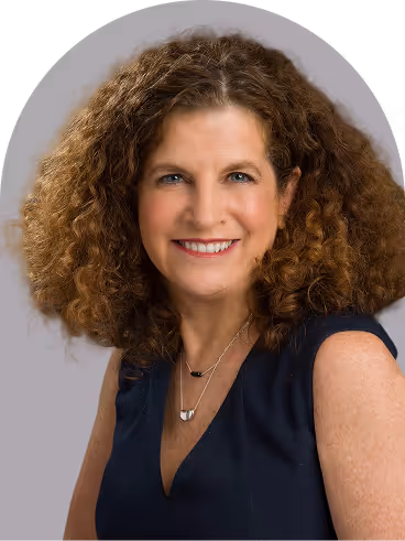 Portrait of a smiling woman with curly brown hair wearing a sleeveless navy top and layered necklaces against a light gray background.