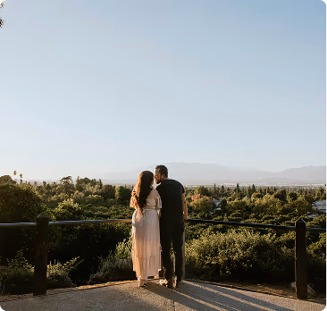 Couple standing close together on a paved lookout, gazing at a scenic view of trees and distant mountains under a clear sky.