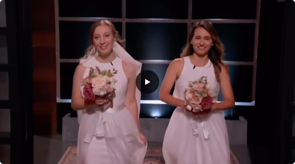 Two smiling women in white dresses holding bouquets, standing side by side indoors.