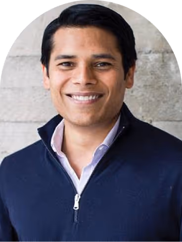 Smiling man with short black hair wearing a navy blue zip-up sweater over a light purple collared shirt, standing in front of a light gray textured wall.