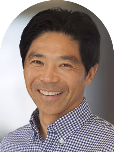 Smiling Asian man wearing a checkered shirt with short black hair against a blurred background.