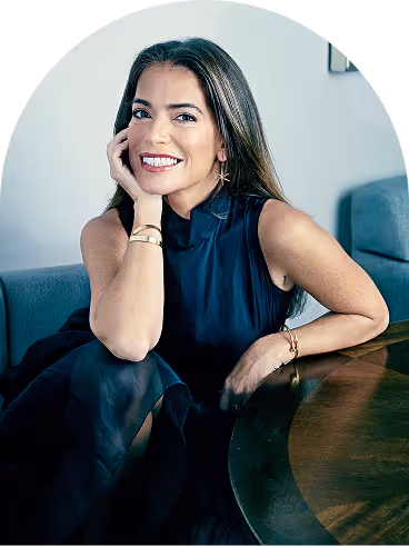 Smiling woman with long brown hair sitting at a wooden table, resting her head on her hand.