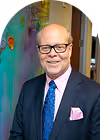 Smiling elderly man wearing glasses, a blue suit, white shirt, blue patterned tie, and pink pocket square, standing indoors near a colorful wall.