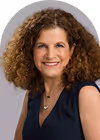 Smiling woman with curly brown hair wearing a navy blue sleeveless top and a delicate necklace.