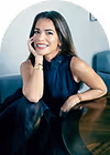 Woman in a sleeveless dark blue dress sitting at a table, smiling with her hand resting on her cheek.
