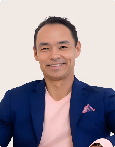 Smiling man wearing a blue blazer with a pink pocket square and light pink shirt against a plain background.