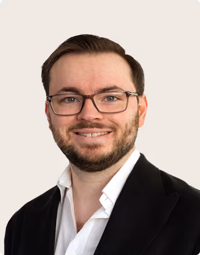Smiling man with glasses, dark hair, and beard wearing a black blazer and white shirt against a light background.