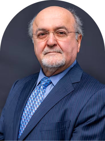 Portrait of an older man with glasses, beard, and mustache wearing a blue suit and patterned tie against a dark background.
