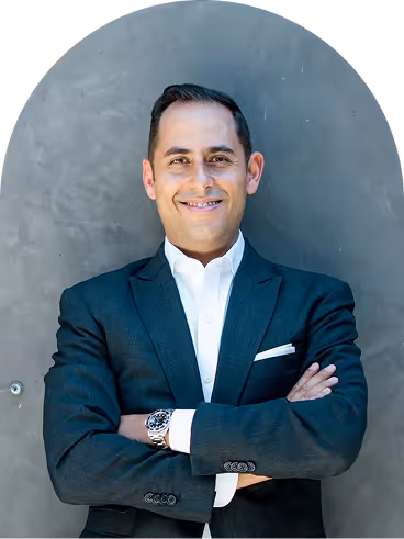 Smiling man in a dark suit with white shirt standing with arms crossed against a gray wall.