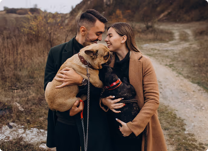 Smiling couple outdoors holding two small dogs close, sharing a joyful moment on a rural path.