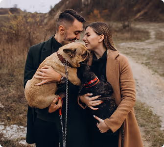 Couple outdoors on a dirt path smiling and holding two French bulldogs.