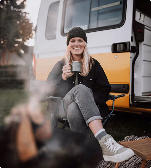Smiling woman in a black beanie and jacket sitting on a camp chair holding a mug, with a yellow camper van in the background.