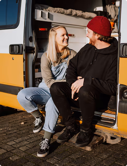 Smiling young woman and man sitting together at the open side door of a yellow camper van.