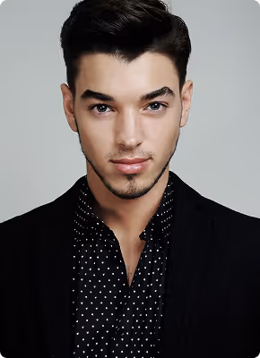 Young man with dark hair wearing a black blazer and black shirt with white polka dots looking confidently at the camera.