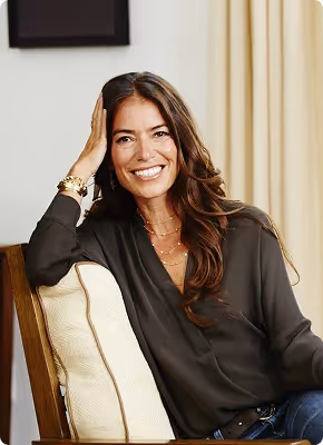 Smiling woman with long brown hair sitting on a chair with a cushion, resting her head on her hand.