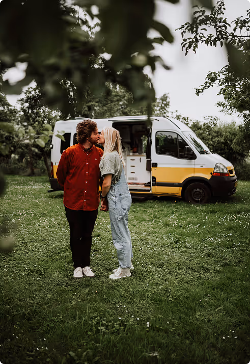 Couple holding hands and kissing outdoors in front of a yellow and white campervan surrounded by greenery.