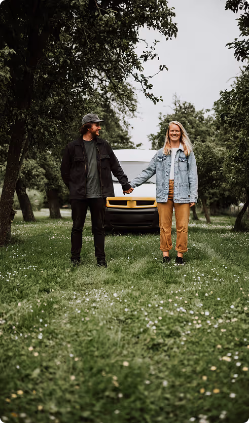 A man and woman holding hands and standing on a grassy path between trees with a white van behind them.