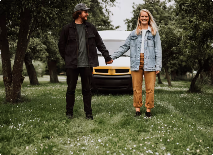 A man and woman holding hands and standing on grass between trees with a white and yellow van behind them.
