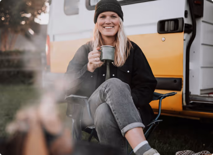Smiling woman in a black beanie and jacket, sitting on a camping chair holding a mug in front of a yellow and white camper van.