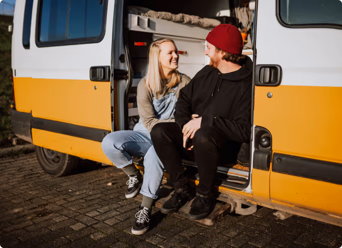A smiling young couple sitting in the open door of a yellow and white camper van, enjoying a sunny day outdoors.