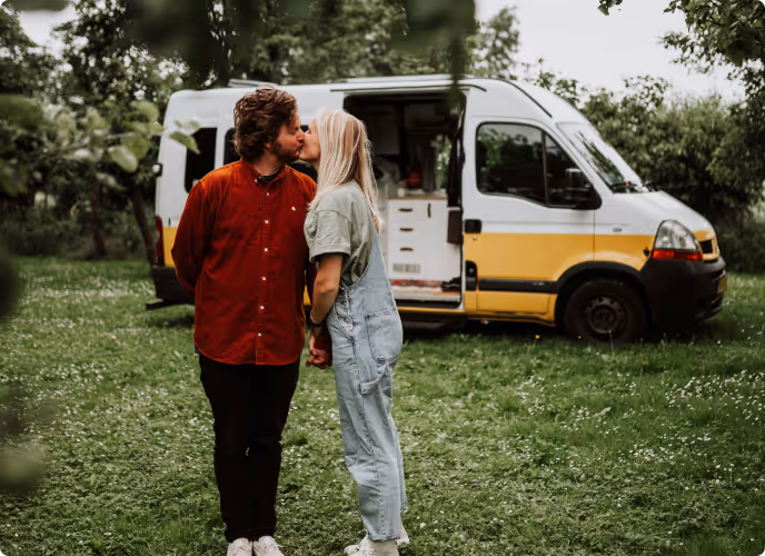Couple kissing while holding hands in front of a white and yellow camper van parked on grass with trees around.