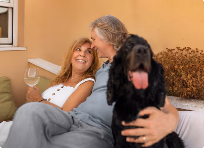Smiling middle-aged couple sitting close together on a couch, holding a black dog with its tongue out and a glass of white wine.