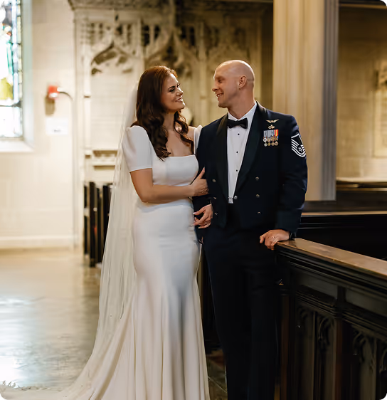 Bride in white gown and veil smiling while holding arm of groom in formal military dress uniform inside a church.