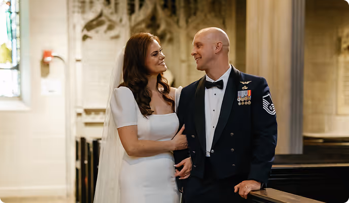 Bride in white dress and groom in military uniform smiling and holding hands inside a church.