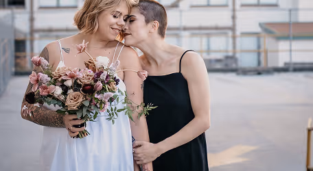 Couple with short hair wearing black and white dresses, holding a bouquet of flowers and sharing a tender moment outdoors.
