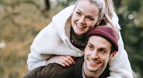 Smiling woman in a white jacket on the back of a smiling man wearing a maroon beanie outdoors.
