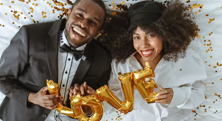 Smiling couple lying down holding gold balloon letters spelling LOVE surrounded by confetti.