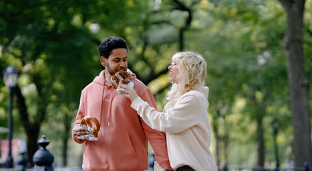Couple in a park smiling as woman offers a pretzel to the man.