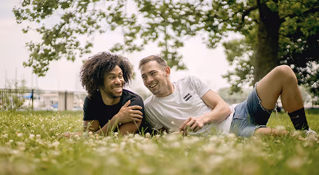 Two young men lying on the grass in a park, smiling and enjoying a sunny day.
