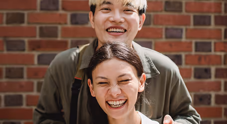 Smiling young man with dyed hair standing behind a laughing young woman against a brick wall.