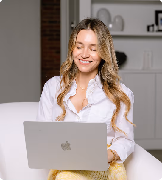 Smiling woman with long blonde hair using a silver Apple laptop while sitting on a white chair in a modern indoor setting.