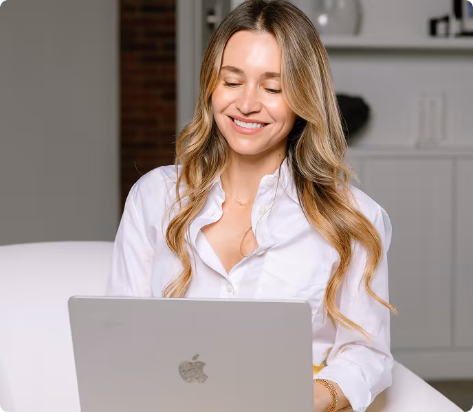Smiling woman with long blonde hair wearing a white blouse, sitting and using a silver Apple laptop.