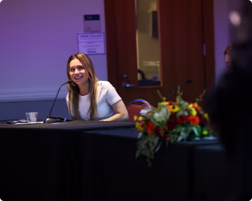 Smiling woman speaking into a microphone while sitting at a conference table with a bouquet of flowers in the foreground.