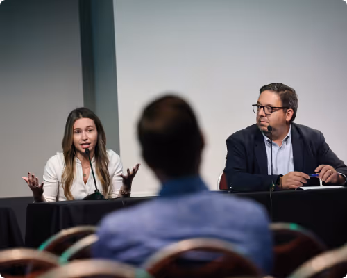 A woman speaking at a panel discussion with a man seated next to her and an audience member in the foreground.