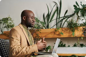 Man in a tan blazer speaking and gesturing while sitting with an open laptop in a plant-filled room.