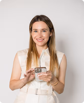 Smiling woman with long hair wearing a sleeveless beige outfit holding a smartphone.